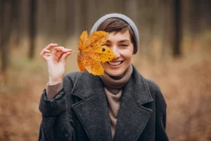 Frau steht im herbstlichen Wald und hält sich lächelnd ein Blatt vor das Gesicht